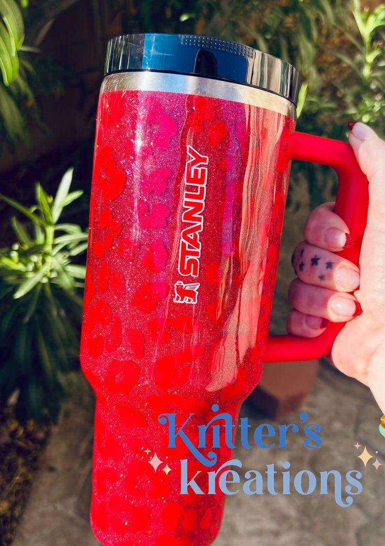 May include: A red Stanley tumbler with a black lid and handle. The tumbler has a glittery leopard print design and the Stanley logo. The text "Kritter's Kreations" is displayed in blue script on the bottom of the tumbler.