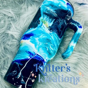 May include: A blue and black stainless steel travel mug with a handle. The mug has a swirling abstract design with blue, white, and black colors. The mug is on a fuzzy gray surface.