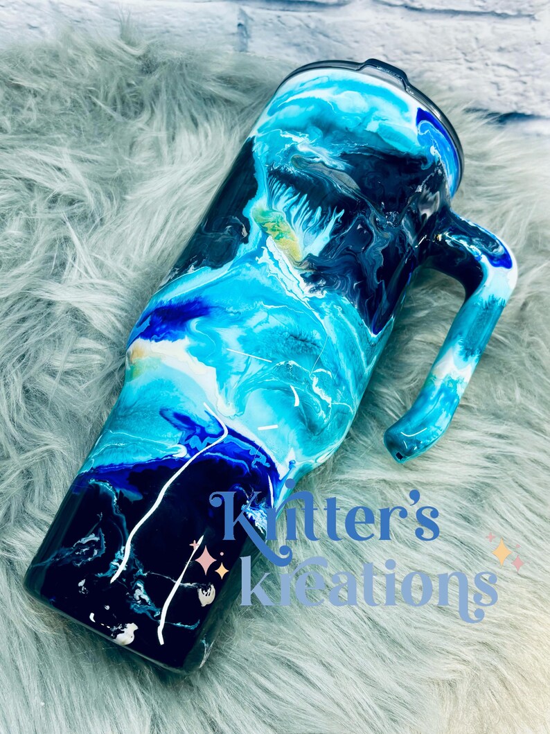 May include: A blue and black stainless steel travel mug with a handle. The mug has a swirling abstract design with blue, white, and black colors. The mug is on a fuzzy gray surface.