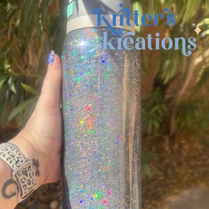 May include: A silver glitter water bottle with a black and gray lid. The bottle has the text "Kritter's Kreations" in light blue. The bottle is cylindrical and appears to be made of stainless steel.