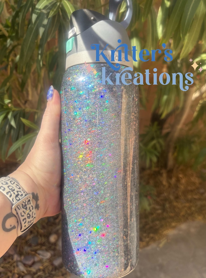 May include: A silver glitter water bottle with a black and gray lid. The bottle has the text "Kritter's Kreations" in light blue. The bottle is cylindrical and appears to be made of stainless steel.