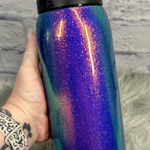 Peut inclure: Une bouteille d'eau cylindrique avec un couvercle en plastique noir dot&eacute; d'un m&eacute;canisme de verrouillage &agrave; bouton-poussoir. Le corps de la bouteille pr&eacute;sente une finition scintillante vibrante aux reflets changeants de violet, bleu et sarcelle.