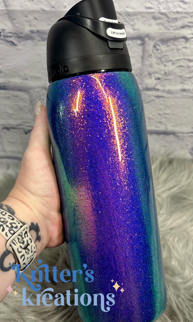 Peut inclure: Une bouteille d'eau cylindrique avec un couvercle en plastique noir dot&eacute; d'un m&eacute;canisme de verrouillage &agrave; bouton-poussoir. Le corps de la bouteille pr&eacute;sente une finition scintillante vibrante aux reflets changeants de violet, bleu et sarcelle.