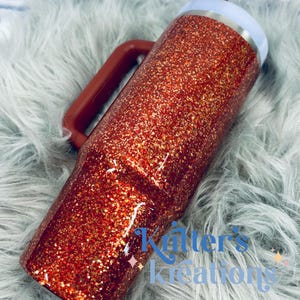 May include: A glitter-covered tumbler in shades of red and gold, featuring a handle and a white rim. The tumbler has the text "Kitter's Kreations" in blue. The handle is a dark red color.