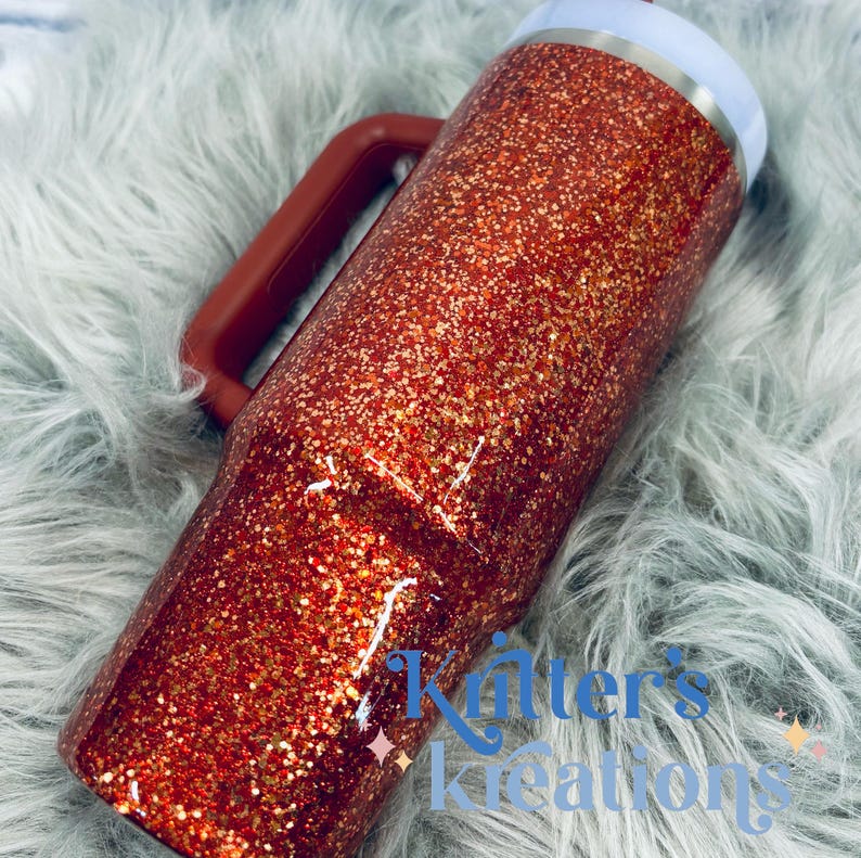 May include: A glitter-covered tumbler in shades of red and gold, featuring a handle and a white rim. The tumbler has the text "Kitter's Kreations" in blue. The handle is a dark red color.