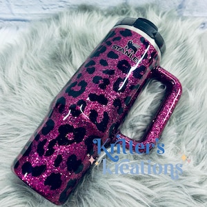 K&ouml;nnte beinhalten: Ein magenta-glitzernder Stanley-Becher mit einem schwarzen Leopardenmuster. Der Becher hat einen Griff und einen schwarzen Deckel. Das Stanley-Logo ist sichtbar. Der Text "Kitter's Creations" befindet sich an der Seite.