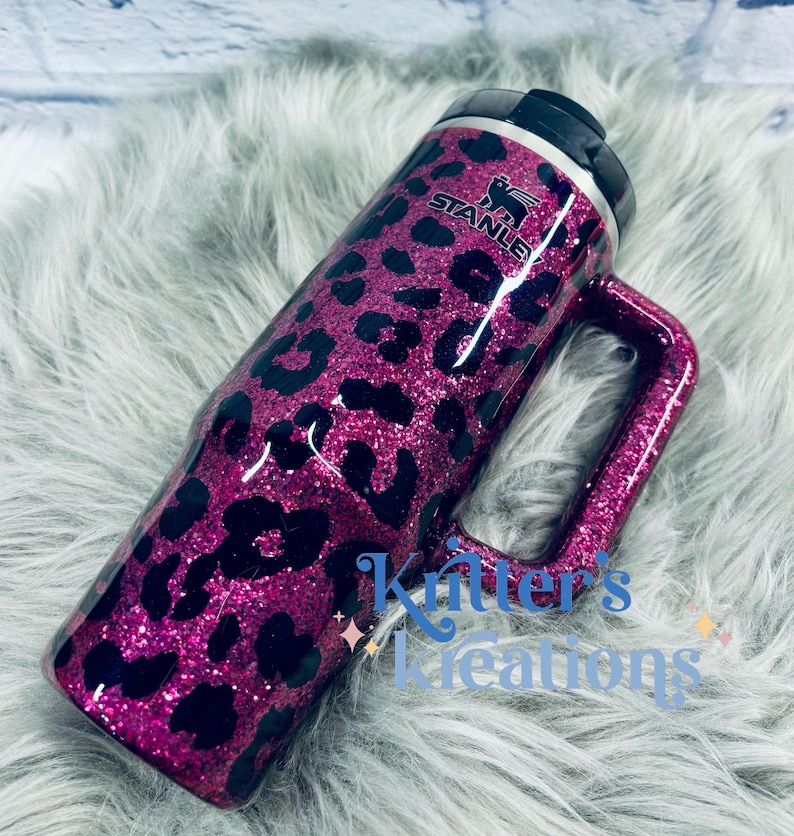 K&ouml;nnte beinhalten: Ein magenta-glitzernder Stanley-Becher mit einem schwarzen Leopardenmuster. Der Becher hat einen Griff und einen schwarzen Deckel. Das Stanley-Logo ist sichtbar. Der Text "Kitter's Creations" befindet sich an der Seite.