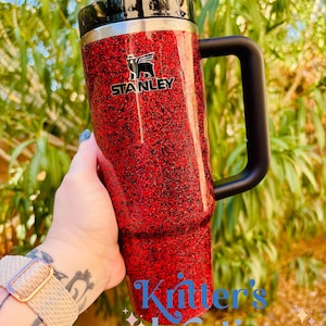 May include: A red glitter Stanley tumbler with a black handle and lid. The Stanley logo is visible on the front. The tumbler is held in front of a green leafy background. The text "Kritter's Kreations" is written in blue cursive.