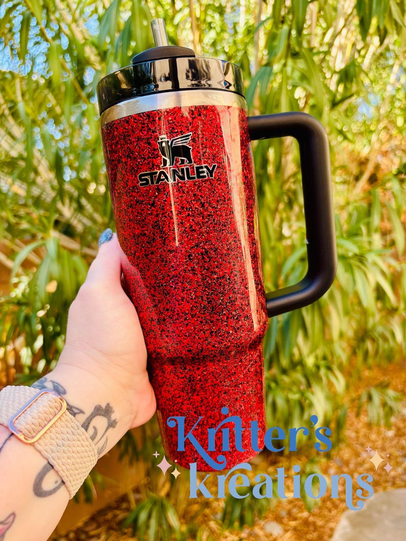 May include: A red glitter Stanley tumbler with a black handle and lid. The Stanley logo is visible on the front. The tumbler is held in front of a green leafy background. The text "Kritter's Kreations" is written in blue cursive.