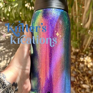 Peut inclure: Une bouteille d'eau cylindrique Owala avec un couvercle noir et une finition &agrave; paillettes holographiques multicolores vibrantes qui alterne entre des tons violets, bleus et verts. Le texte "Knitter's kreations" est imprim&eacute; en bleu sur le c&ocirc;t&eacute;.