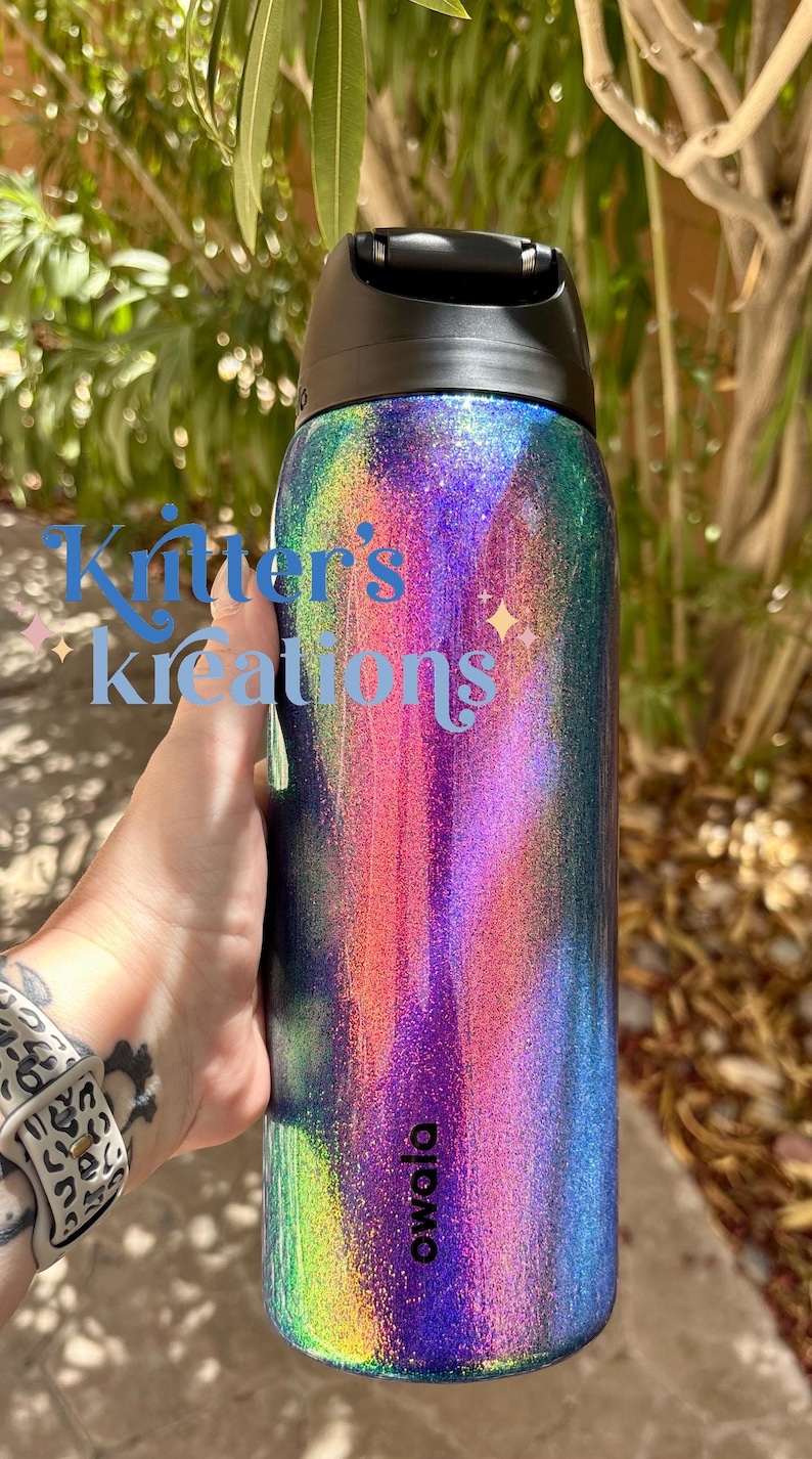 Peut inclure: Une bouteille d'eau cylindrique Owala avec un couvercle noir et une finition &agrave; paillettes holographiques multicolores vibrantes qui alterne entre des tons violets, bleus et verts. Le texte "Knitter's kreations" est imprim&eacute; en bleu sur le c&ocirc;t&eacute;.