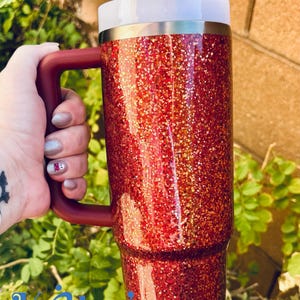 May include: A red glitter tumbler with a matching handle and lid. The tumbler has a white rim and a straw. The handle is a dark red color. The tumbler is held by a hand.