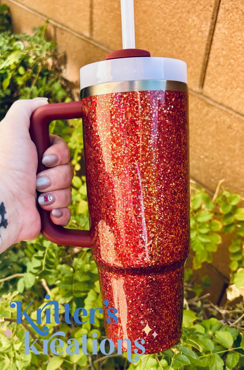 May include: A red glitter tumbler with a matching handle and lid. The tumbler has a white rim and a straw. The handle is a dark red color. The tumbler is held by a hand.