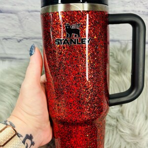 May include: A red glitter-covered Stanley tumbler with a black handle and lid. The Stanley logo is visible on the front. The tumbler has a straw and the text "Kritter's Kreations" is at the bottom.