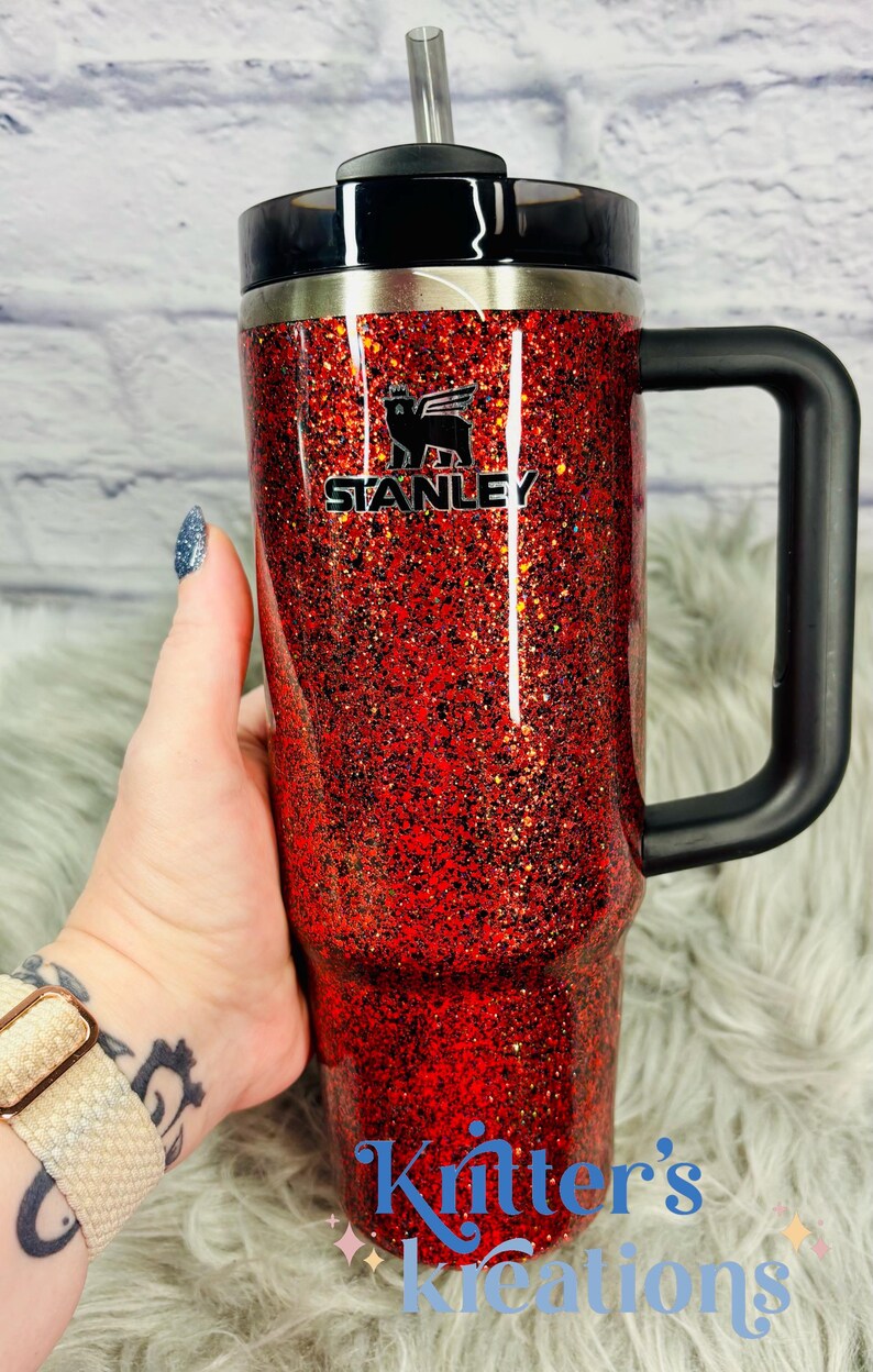May include: A red glitter-covered Stanley tumbler with a black handle and lid. The Stanley logo is visible on the front. The tumbler has a straw and the text "Kritter's Kreations" is at the bottom.