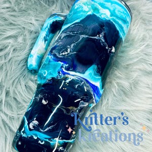 May include: A black and blue tumbler with a white handle. The tumbler has a marbled design with swirls of blue, black, and white. The tumbler is on a light blue fuzzy surface. The text "Knitter's Kreations" is visible in the bottom right corner of the image.
