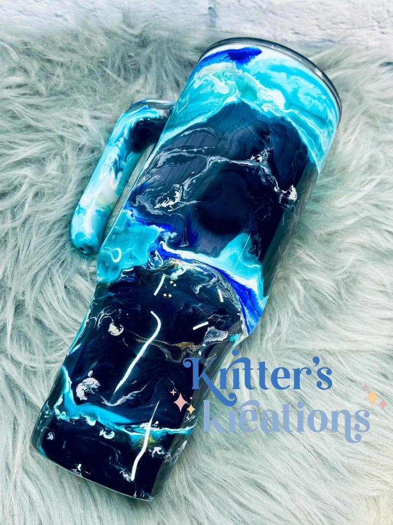 May include: A black and blue tumbler with a white handle. The tumbler has a marbled design with swirls of blue, black, and white. The tumbler is on a light blue fuzzy surface. The text "Knitter's Kreations" is visible in the bottom right corner of the image.