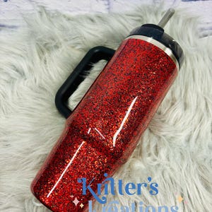 May include: A red glitter tumbler with a black handle and lid. The tumbler has a silver straw and the text "Kitter's Kreations" in blue. The tumbler is on a white fluffy surface.
