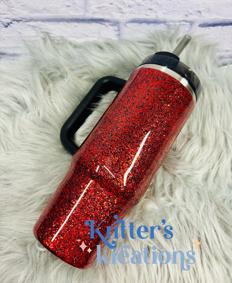 May include: A red glitter tumbler with a black handle and lid. The tumbler has a silver straw and the text "Kitter's Kreations" in blue. The tumbler is on a white fluffy surface.