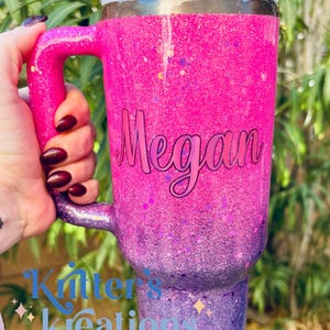 May include: A large, glitter-covered tumbler in shades of pink and purple, with a handle and a lid. The name "Megan" is written on the side in cursive. The tumbler has a straw and is held by a person.