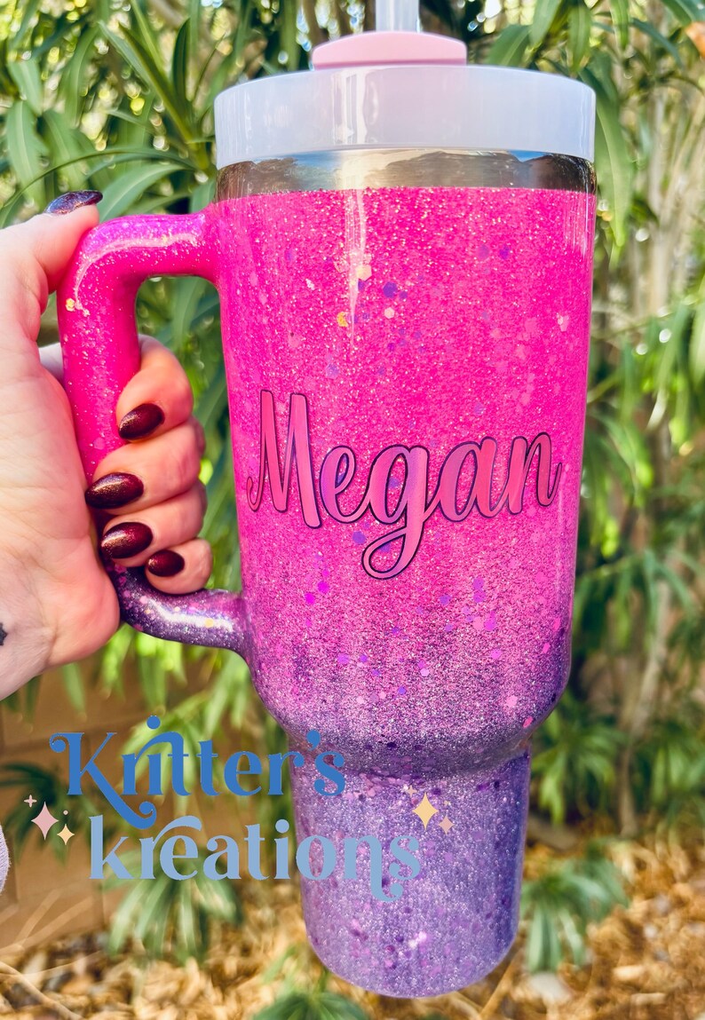 May include: A large, glitter-covered tumbler in shades of pink and purple, with a handle and a lid. The name "Megan" is written on the side in cursive. The tumbler has a straw and is held by a person.