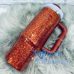 May include: A glitter-covered Stanley tumbler in shades of orange and gold. The tumbler has a handle and a white and red lid. The Stanley logo is visible on the side. The text "Kitter's Kreations" is written on the bottom of the tumbler.