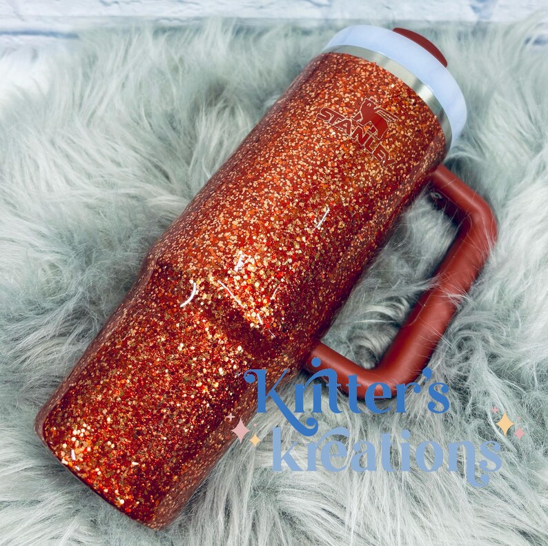 May include: A glitter-covered Stanley tumbler in shades of orange and gold. The tumbler has a handle and a white and red lid. The Stanley logo is visible on the side. The text "Kitter's Kreations" is written on the bottom of the tumbler.