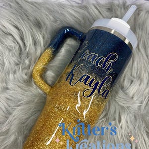 Puede incluir: Vaso con purpurina y asa, con un dise&ntilde;o degradado azul marino y dorado. El vaso tiene el nombre "Coach Kayla" en escritura azul marino y el texto "Killer's Kreations" en azul. Incluye tapa y pajita blancas.