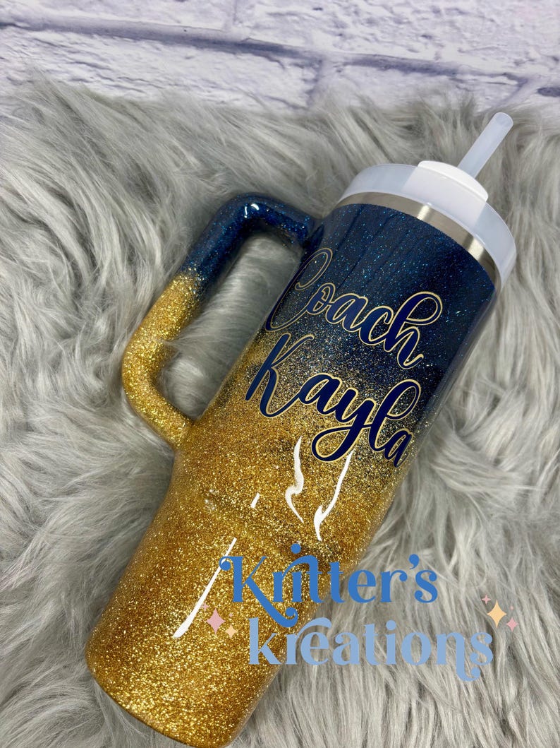 Puede incluir: Vaso con purpurina y asa, con un dise&ntilde;o degradado azul marino y dorado. El vaso tiene el nombre "Coach Kayla" en escritura azul marino y el texto "Killer's Kreations" en azul. Incluye tapa y pajita blancas.