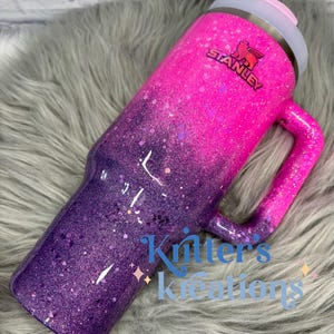 May include: A glittery Stanley tumbler in a gradient of pink and purple. The tumbler has a handle and a lid with a pink slider. The Stanley logo is visible. The text "Kritter's Kreations" is also visible.
