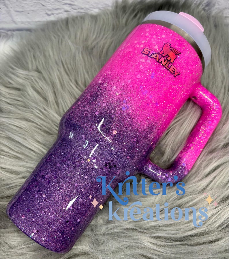 May include: A glittery Stanley tumbler in a gradient of pink and purple. The tumbler has a handle and a lid with a pink slider. The Stanley logo is visible. The text "Kritter's Kreations" is also visible.