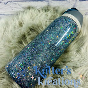 May include: A glitter-covered Owala water bottle. The bottle is primarily blue with iridescent glitter, and has a gray lid. The text "Kritter's Kreations" is visible on the bottle. The bottle is resting on a fluffy, light gray surface.