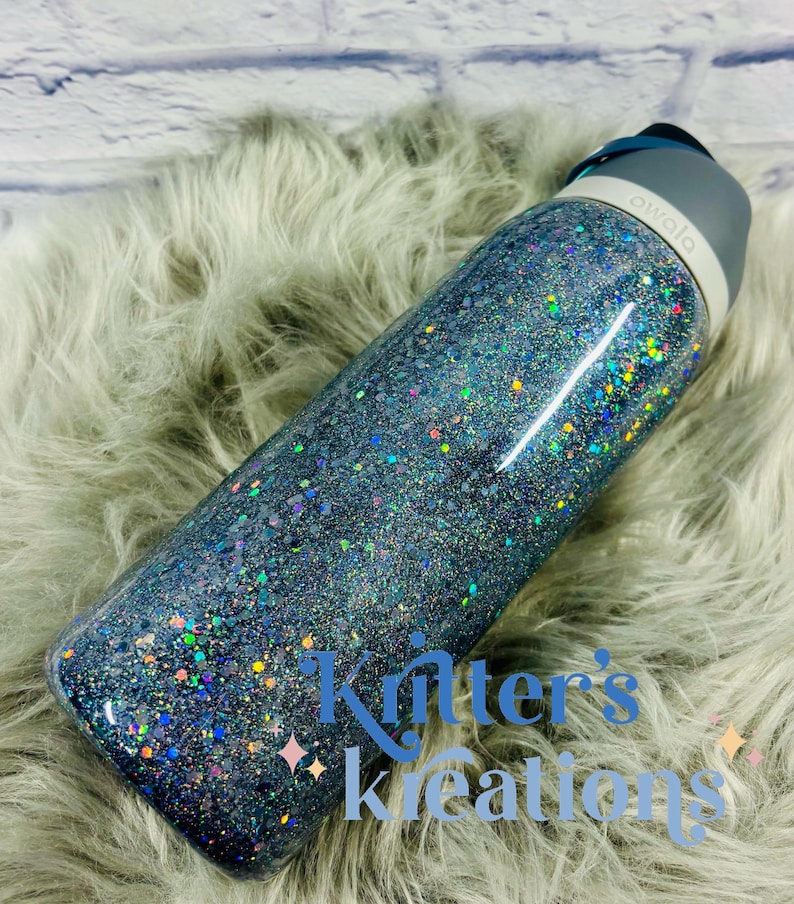 May include: A glitter-covered Owala water bottle. The bottle is primarily blue with iridescent glitter, and has a gray lid. The text "Kritter's Kreations" is visible on the bottle. The bottle is resting on a fluffy, light gray surface.