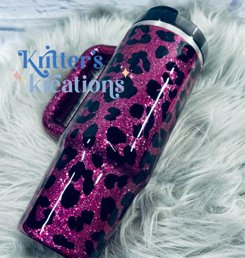K&ouml;nnte beinhalten: Ein glitzernder, pinkfarbener Becher mit einem schwarzen Leopardenmuster. Der Becher hat einen Griff und einen schwarzen Deckel. Der Text "Kritter's Kreations" ist auf der Seite des Bechers zu sehen.