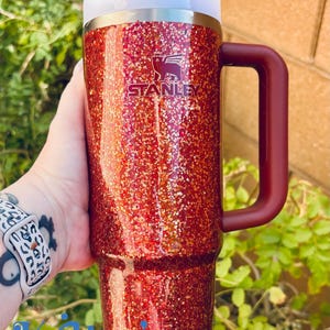 May include: A red glitter Stanley tumbler with a white lid and a burgundy handle. The Stanley logo is visible on the front. The tumbler is held in front of a green leafy background. The text "Kritter's Kreations" is at the bottom.