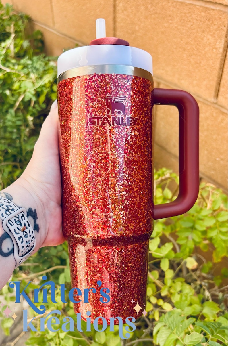 May include: A red glitter Stanley tumbler with a white lid and a burgundy handle. The Stanley logo is visible on the front. The tumbler is held in front of a green leafy background. The text "Kritter's Kreations" is at the bottom.