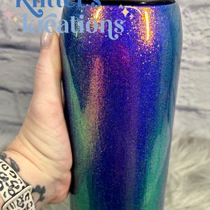 Peut inclure: Une bouteille d'eau cylindrique Owala avec un couvercle noir et une finition paillet&eacute;e passant du bleu profond au violet et au vert. Le texte Kitter's Kreations est imprim&eacute; en bleu clair sur la partie sup&eacute;rieure et Owala est imprim&eacute; en noir.