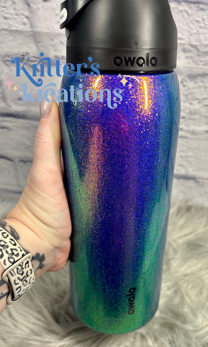 Peut inclure: Une bouteille d'eau cylindrique Owala avec un couvercle noir et une finition paillet&eacute;e passant du bleu profond au violet et au vert. Le texte Kitter's Kreations est imprim&eacute; en bleu clair sur la partie sup&eacute;rieure et Owala est imprim&eacute; en noir.