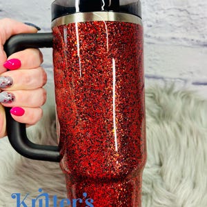 May include: A red glitter tumbler with a black handle, lid, and straw. The tumbler has the text "Kritter's Kreations" in blue. The tumbler is on a fluffy, white surface.