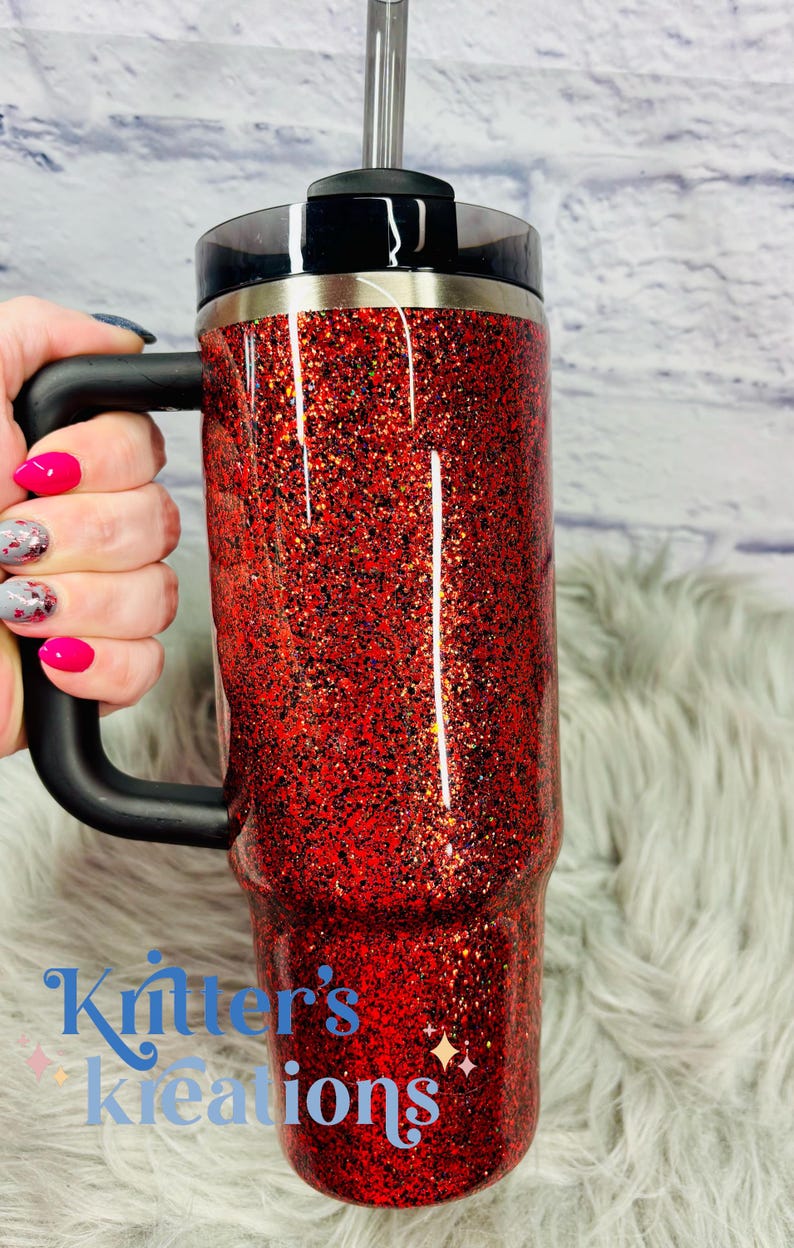 May include: A red glitter tumbler with a black handle, lid, and straw. The tumbler has the text "Kritter's Kreations" in blue. The tumbler is on a fluffy, white surface.