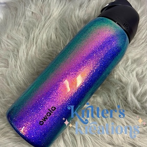 Peut inclure: Une bouteille d'eau cylindrique Owala avec une finition d&eacute;grad&eacute;e scintillante et vibrante passant du violet profond au magenta vif et au bleu sarcelle. La bouteille poss&egrave;de un bouchon noir et le nom de la marque Owala imprim&eacute; en noir.