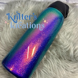 Peut inclure: Une bouteille d'eau cylindrique avec un couvercle noir et une finition scintillante passant par des nuances de bleu profond, de violet vibrant et de vert sarcelle. Le texte "Kritter's Kreations" est imprim&eacute; en bleu sur le c&ocirc;t&eacute;.