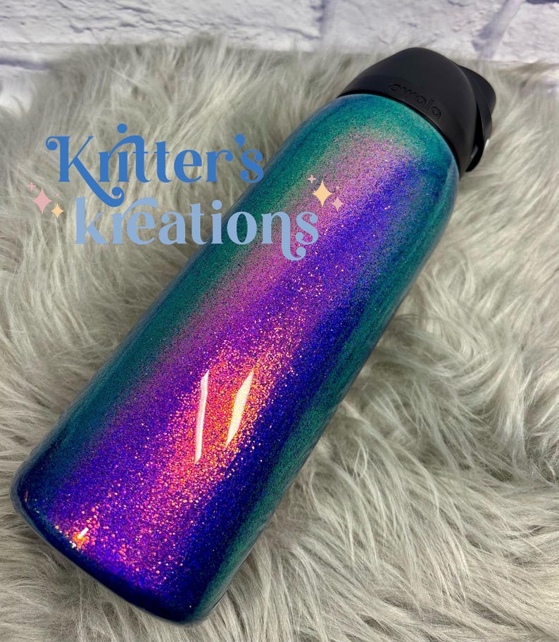 Peut inclure: Une bouteille d'eau cylindrique avec un couvercle noir et une finition scintillante passant par des nuances de bleu profond, de violet vibrant et de vert sarcelle. Le texte "Kritter's Kreations" est imprim&eacute; en bleu sur le c&ocirc;t&eacute;.