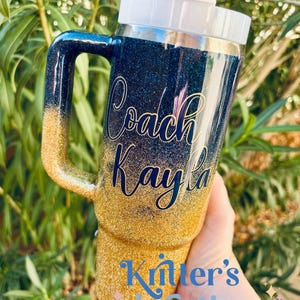 Puede incluir: Vaso cubierto de purpurina con asa, con un degradado de oro a azul marino. El texto "Coach Kayla" est&aacute; en cursiva. La parte inferior del vaso tiene el texto "Knitter's Kreations". Se ve una pajita y una tapa blancas.