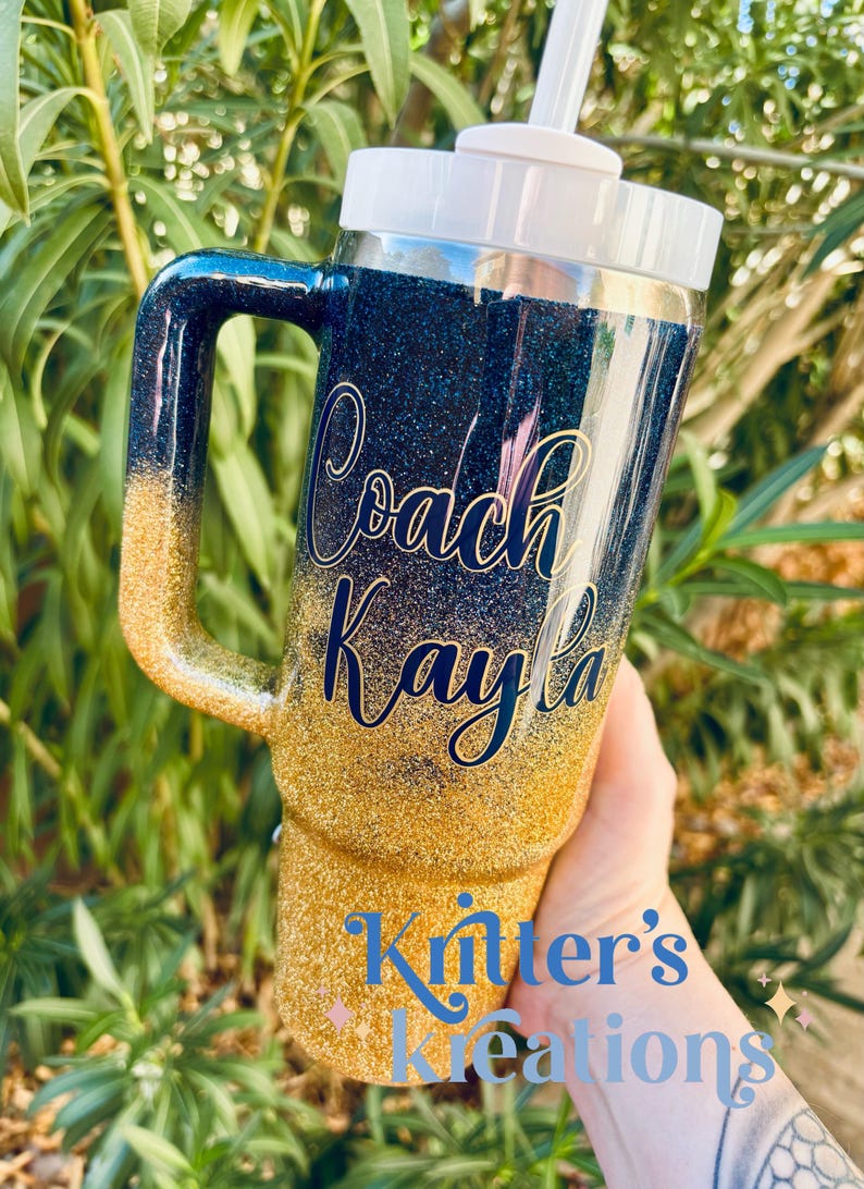 Puede incluir: Vaso cubierto de purpurina con asa, con un degradado de oro a azul marino. El texto "Coach Kayla" est&aacute; en cursiva. La parte inferior del vaso tiene el texto "Knitter's Kreations". Se ve una pajita y una tapa blancas.