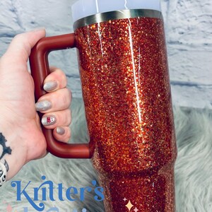 May include: A red glitter tumbler with a handle, a white lid, and a straw. The tumbler has a metallic silver band near the top. The handle and the top of the lid are the same shade of red. The text "Kritter's Kreations" is visible in the image.