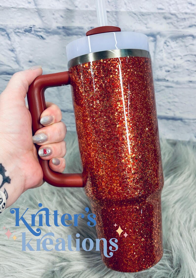 May include: A red glitter tumbler with a handle, a white lid, and a straw. The tumbler has a metallic silver band near the top. The handle and the top of the lid are the same shade of red. The text "Kritter's Kreations" is visible in the image.