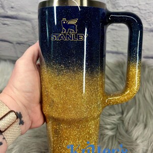 Puede incluir: Un vaso Stanley con asa, con un dise&ntilde;o degradado de purpurina azul marino a dorado. El vaso tiene una tapa blanca y una pajita. El logotipo de Stanley es visible en dorado. El texto "Killer's Kreations" est&aacute; en la parte inferior.