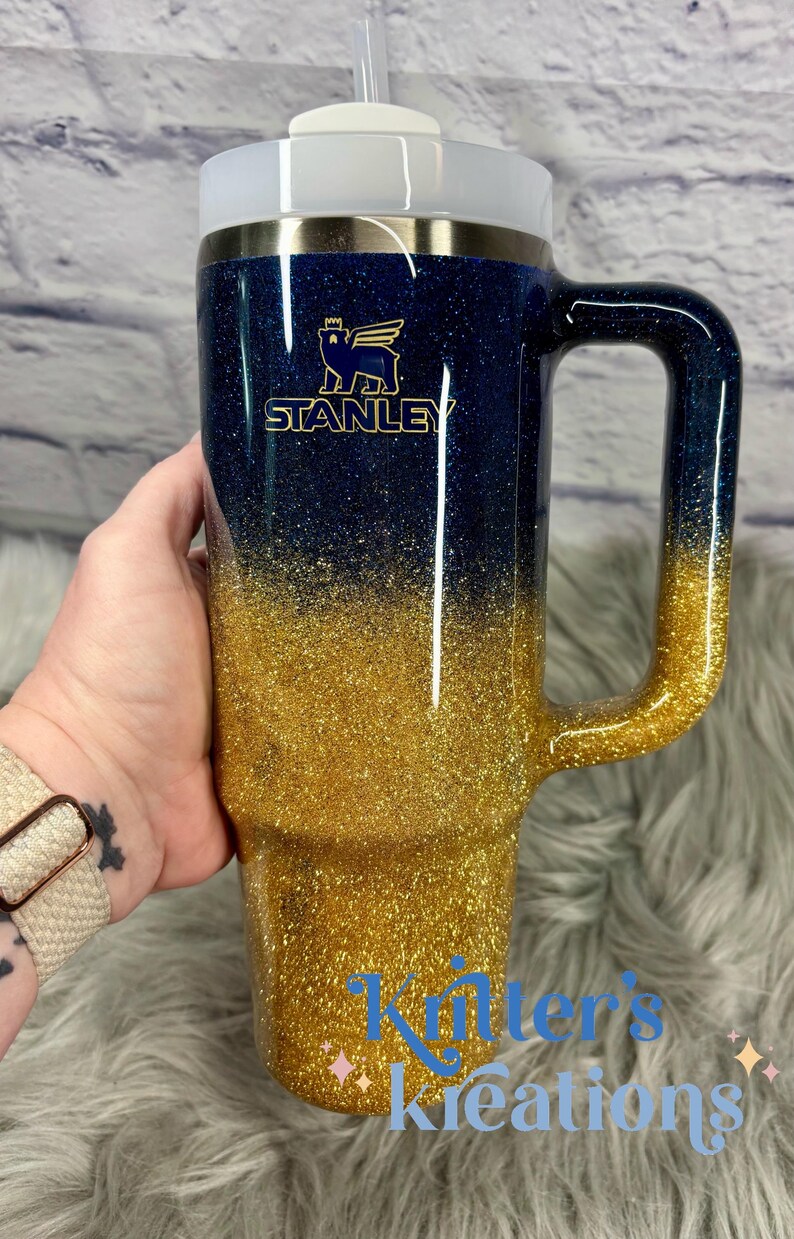 Puede incluir: Un vaso Stanley con asa, con un dise&ntilde;o degradado de purpurina azul marino a dorado. El vaso tiene una tapa blanca y una pajita. El logotipo de Stanley es visible en dorado. El texto "Killer's Kreations" est&aacute; en la parte inferior.