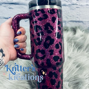 K&ouml;nnte beinhalten: Ein pinkfarbener Reisebecher mit Glitzer, schwarzem Deckel und Griff. Der Becher ist mit einem schwarzen Leopardenmuster versehen. Der Text "Kritter's Kreations" ist auf der Seite des Bechers zu sehen.
